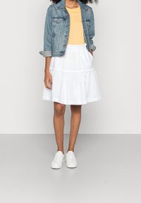 Džínová bunda, žluté tričko a bílá sukni ve vrstvách, doplněné bílými teniskami. Tento outfit se vyznačuje pohodlným a vrstveným designem.