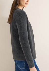 Šedý chlupatý svetr s texturovaným povrchem, kulatým výstřihem a dlouhými rukávy, ve spojení s modrými džínami na neutrálním pozadí.