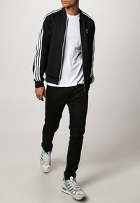 Black track jacket with white three-stripe design, white t-shirt, black pants, and gray sneakers with green accents.