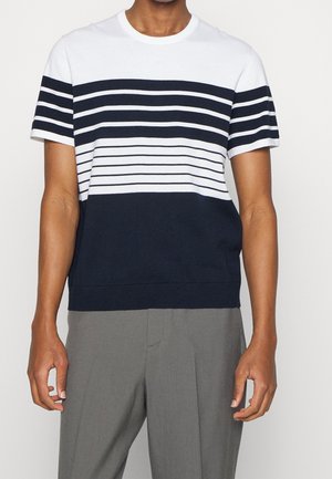 Person trägt ein weißes und marineblaues kurzärmeliges Hemd mit horizontalen Streifen und graue Hose, steht vor einem schlichten Hintergrund.