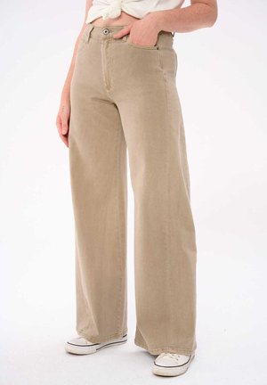 Osoba ubrana w beżowe szerokie spodnie jeansowe, białe trampki i białą włożoną do środka koszulkę, stojąca z jedną ręką w kieszeni na białym tle.