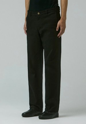 Mann trägt lockere schwarze Hose, schwarze Schuhe und ein schwarzes ärmelloses Oberteil, steht vor einem einfarbigen hellgrauen Hintergrund.