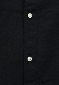 Μαύρο ύφασμα με δύο ορατά λευκά κουμπιά ραμμένα κάθετα, δείχνοντας λεπτομερή ράψιμο κατά μήκος της λωρίδας κουμπιών.