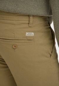 Beige katoenen broek met een achterzak voorzien van een houten knoop. Een klein label met de tekst "DEDICATED TO DENIM PERFECTION." Eenvoudig, strak ontwerp.