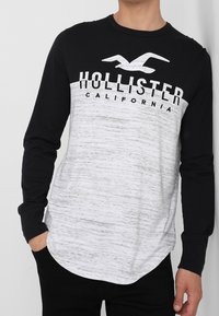 Pitkähihaine t-paita, jossa on musta yläosa, valkoinen alaosa, joka sisältää tekstuuripinnan, ja "HOLLISTER CALIFORNIA" -teksti painettuna rintaan.