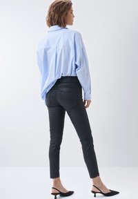 Camisa de rayas azul y blanca de mangas largas, jeans ajustados negros y mules negros con tacón, vistas de lado y por la parte de atrás sobre un fondo blanco.