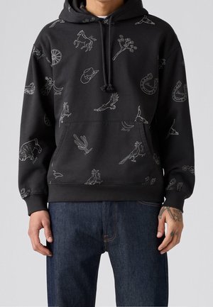Mężczyzna w czarnej bluzie z kapturem z białymi konturami ilustracji w stylu western i niebieskich dżinsach, stojący na szarym tle.