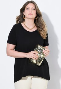 Top nero a maniche corte con scollatura a barca, abbinato a pantaloni chiari con dettagli decorativi. Tiene in mano una pochette dorata con paillettes.