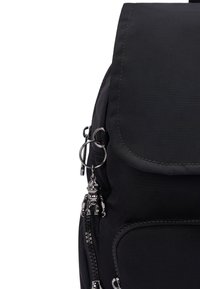 Black nylon backpack with a flap closure, front zip pocket, and a metallic toy keychain attached. Smooth texture and minimalist design.