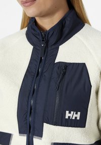 Chaqueta de forro polar con un cuerpo suave en color crema y detalles en azul marino, un cuello alto y bolsillos con cremallera. Logo notable en el pecho.
