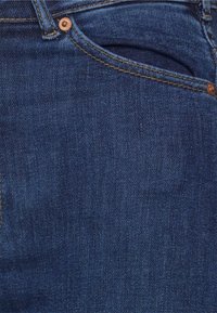 Lähikuva tummansinisistä farkuista, joissa on etutasku, metalliset niitit ja näkyvät saumat.