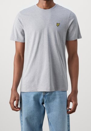 Graues Baumwoll-T-Shirt mit kurzen Ärmeln, rundem Halsausschnitt und gelbem, besticktem Logo auf der linken Brust. Kombiniert mit hellblauen Jeans.