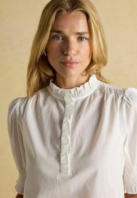 Blonde Frau mit blauen Augen, die eine weiße Bluse mit Rüschensoller und Knopfleiste vor einem beigen Hintergrund trägt.