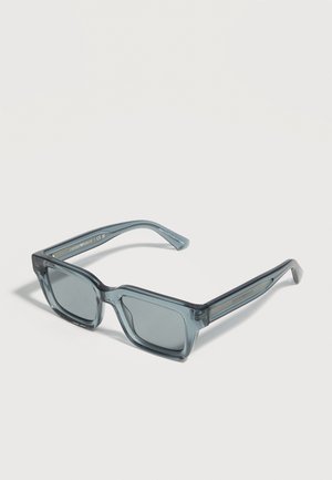 Lunettes de soleil rectangulaires gris transparent avec verres opaques et montures épaisses posées sur une surface blanche.