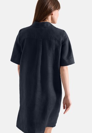 Kvinde med glat brunt hår iført en løs, kortærmet, knælængde sort kjole, set bagfra på en hvid baggrund.