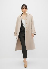 Cappotto di lana beige con colletto, chiusura a bottoni e tasche laterali, indossato sopra un completo nero a righe sottili e décolleté nude. Design semplice e strutturato.