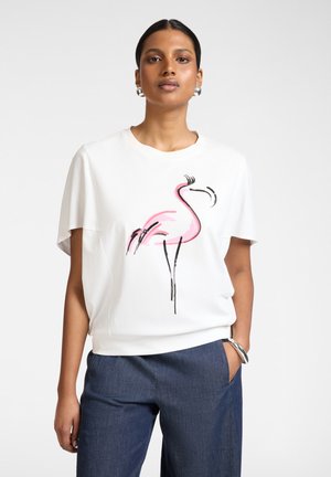 Frau trägt weißes Kurzarmshirt mit pinkem Flamingo-Motiv, blaue Hose, silberne Creolen und silbernes Armband, steht mit einer Hand in der Tasche.