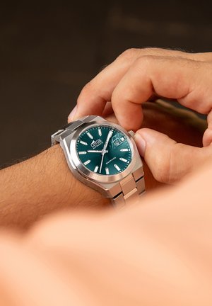 Reloj de acero inoxidable plateado con una esfera de color verde oscuro, marcadores de hora luminosos y una ventana de fecha. Cristal de zafiro sobre la carátula.