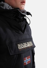 Gros plan d'une personne portant une veste noire Napapijri avec un écusson du drapeau norvégien et des cordons de serrage sur la capuche.