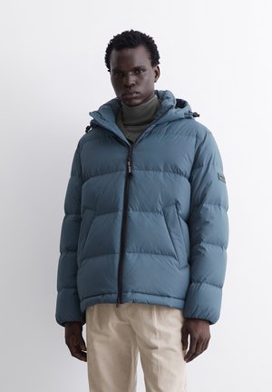 Teal puffer jacket with a zip front, adjustable hood, and stitched detailing. Worn over a dark turtleneck and light beige corduroy pants.