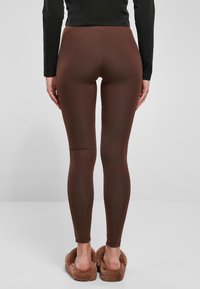 Leggings marron en matière lisse et extensible, présentant une coupe ajustée avec un détail de couture sur le bas de la jambe, portés avec des chaussons duveteux.