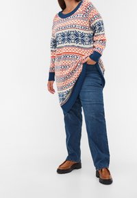 Person klädd i en mönstrad långtröja med blå, orange och vita mönster, blå jeans och bruna klumpiga skor, står med handen i fickan.