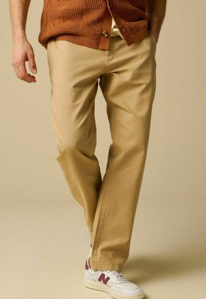 Homme portant un pantalon beige, des baskets blanches avec des accents bordeaux, et un cardigan en tricot marron boutonné, debout devant un fond beige.