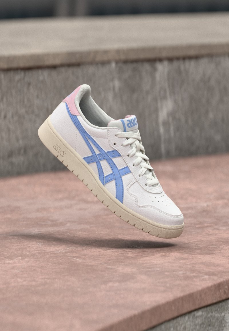 Adidași albi cu dungi albastre și accente roz plutind deasupra unei suprafețe de piatră, evidențiind detalii ale tălpii și șireturilor.