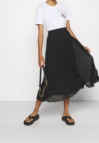 Vrouw in een witte graphic t-shirt, zwarte geplooide midi-rok, zwarte platform flip-flops, en een zwarte handtas met een gouden ketting.