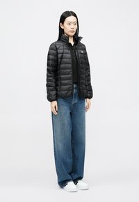 Melna pūtīga jaka ar šūtu dizainu, augstu apkakli, priekšējo rāvējslēdzēju, kopā ar brīvā stila zilām džinsa biksēm un baltām sporta kurpēm.