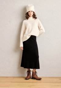 Maglione a costine crema, gonna nera svasata e stivaletti lacciati marroni con rifiniture in pelliccia. Modella posa su uno sfondo neutro.
