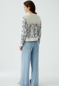 Gestrickter Pullover mit Schlangenmuster in Grau und Weiß, mit einem Mesh-Rücken und einem hellrosa horizontalen Streifen. Kombiniert mit weit geschnittenen blauen Jeans.