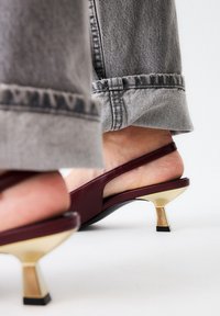Zapatos de tacón estilo slingback de cuero burdeos con detalles en forma geométrica en tono dorado en el tacón. La suela es negra y el zapato tiene un acabado suave.