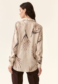 Camisa de manga larga beige con un patrón de hojas en marrón y negro. Tejido suave con cuello tipo polo y puños con botones.