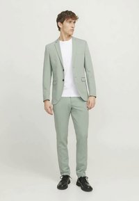 Νεαρός άνδρας που φοράει ένα ανοιχτό πράσινο κοστούμι, λευκή μπλούζα και μαύρες επίσημες παπούτσια, στέκεται και κοιτάζει πλαγίως σε έναν απλό φόντο.