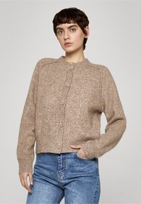 Beige strikket cardigan med rund hals, ribbestrikkede ermekanter og knapper foran. Teksturert stoff med myk følelse, kombinert med høytlivsjeans.