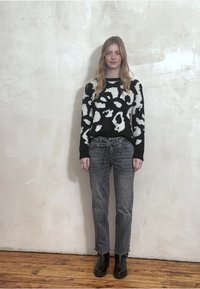 Czarny sweter z białym abstrakcyjnym wzorem, szare jeansy z wiązanym w pasie ściągaczem oraz czarne botki za kostkę. Tło stanowi neutralna, teksturowana ściana.