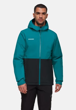 Man draagt een Mammut-jas met capuchon in turquoise en zwart, staand met het gezicht naar voren en ontspannen handen langs zijn zij.
