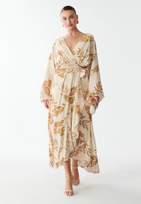 Wikkeljurk in lichtbeige met een goudbladerenpatroon, lange mouwen en een strikceintuur in de taille. Afgemaakt met gouden sandalen met banden.