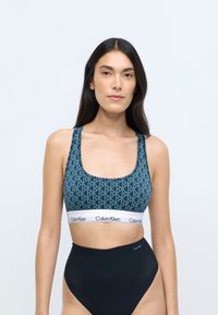 Sportsbh med blåt geometrisk mønster, brede stropper og et hvidt Calvin Klein-logo bånd. Højtaljede sorte trusser komplimenterer designet.