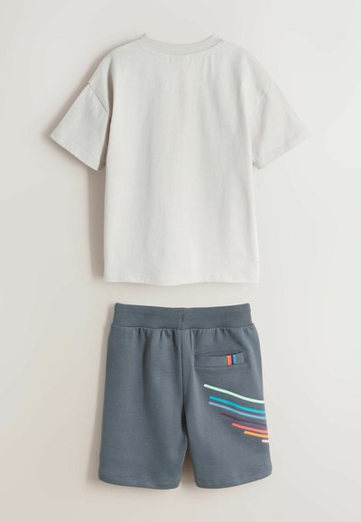 Hvid t-shirt med korte ærmer kombineret med grå shorts med elastisk taljebånd, der har en baglomme og farverige diagonale striber på det ene ben.