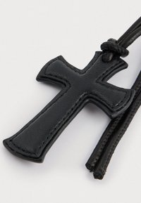 Crna kožna križna privjeska s glatkom teksturom i uzdignutim rubovima, pričvršćena na crnu pletenu uzicu.