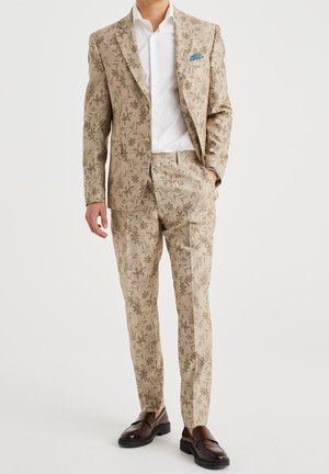 Homme portant un costume beige à motif floral, une chemise blanche, un mouchoir de poche bleu et des mocassins marron foncé, debout avec une main dans la poche.