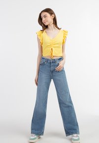 Gul cropped top med korte volangærmer og samlet frontdetalje, parret med bløde blå jeans. Model iført multifarvede sko.