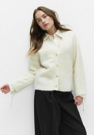 Vit, mjuk ullcardigan med V-hals, knappframsida och långa ärmar. Har en avslappnad passform och knytning vid ärmarna. Svarta vida byxor.