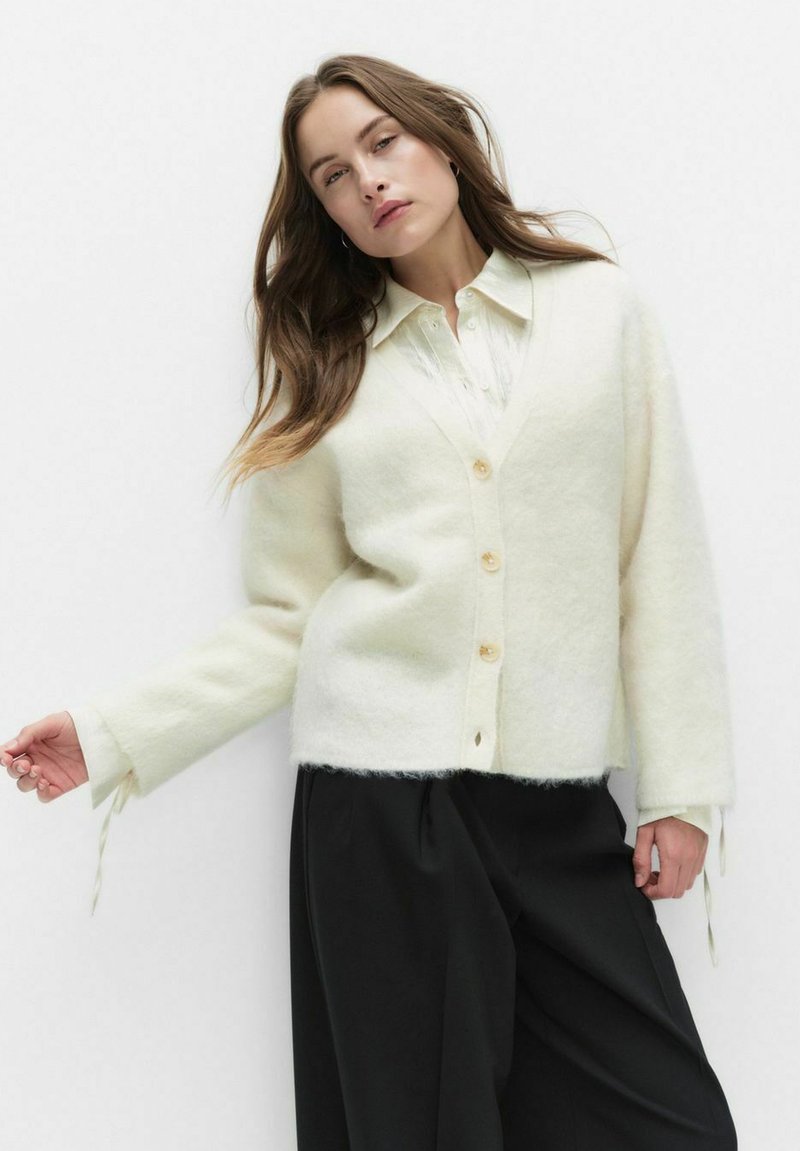 Biały, miękki kardigan wełniany z dekoltem w kształcie litery V, zapinany na guziki, z długimi rękawami. Posiada luźny krój oraz wiązania przy rękawach. Czarne spodnie o szerokich nogawkach.