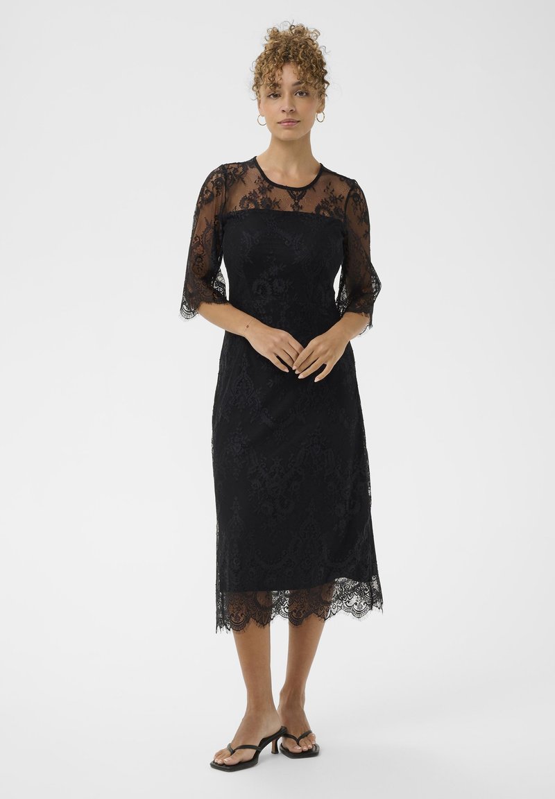 Mujer con cabello rizado vestida con un vestido midi de encaje negro con mangas y escote transparente, de pie con las manos entrelazadas, sobre fondo blanco.