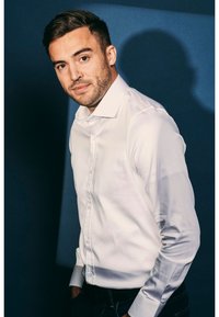 Witte blouse met knopen, met een gladde textuur, met lange mouwen, gecombineerd met donkere jeans tegen een blauwe achtergrond.