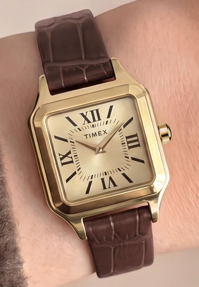 Τετράγωνο ρολόι χειρός Timex σε χρυσή απόχρωση με ρωμαϊκούς αριθμούς και καφέ δερμάτινο λουράκι σε καρπό που δείχνει 11:08.