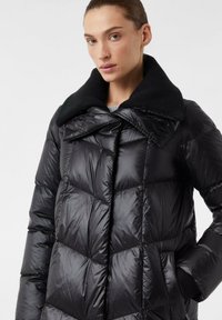 Donna con un giubbotto imbottito nero a trapunta, con un ampio colletto e le mani in tasca, su uno sfondo chiaro e semplice.